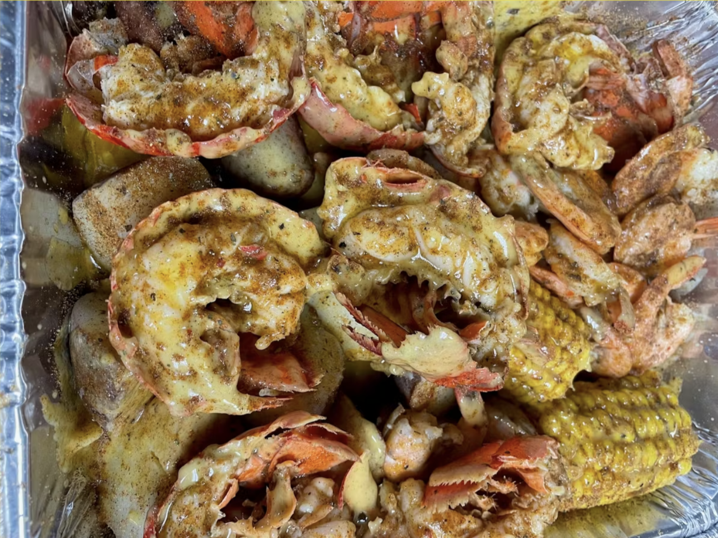A tray of seasoned seafood including lobsters, shrimp, and corn on the cob, all covered in spices, perfect for late night bites.